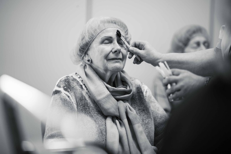 Eine sitzende Patientin mit OP Haube und geschlossenen Augen wird auf den Eingriff vorbereitet. Eine Mitarbeiterin reinigt und desinfiziert den rechten Gesichtsbereich mit zwei gro&szlig;en Wattest&auml;bchen / &copy; Till Findl